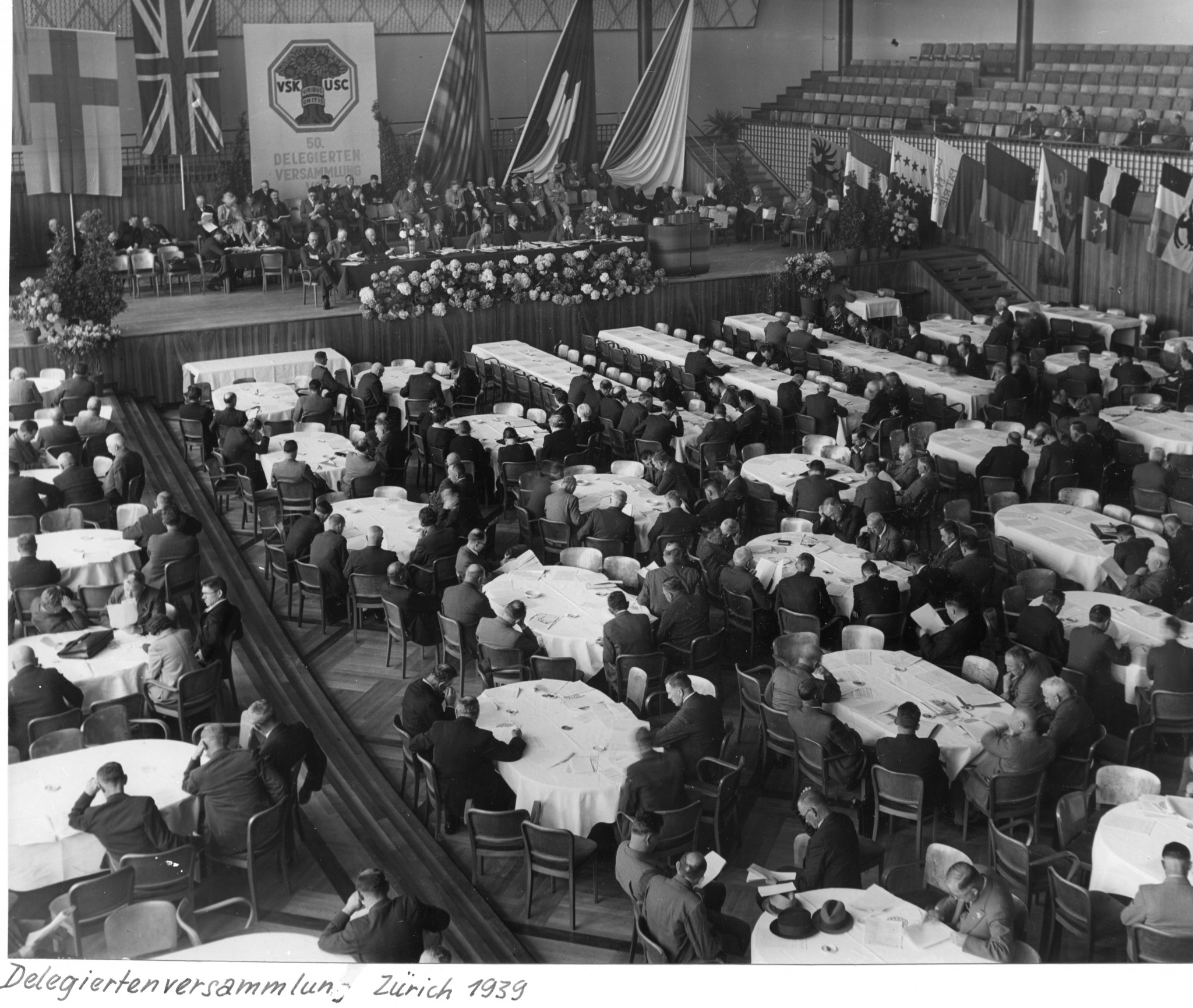 Delegiertenversammlung des Verbandes Schweizerischer Konsumvereine, Zürich 1939: Sitzungssaal mit Vorstand (?) auf der Bühne und Delegierten an Tischen im Saal