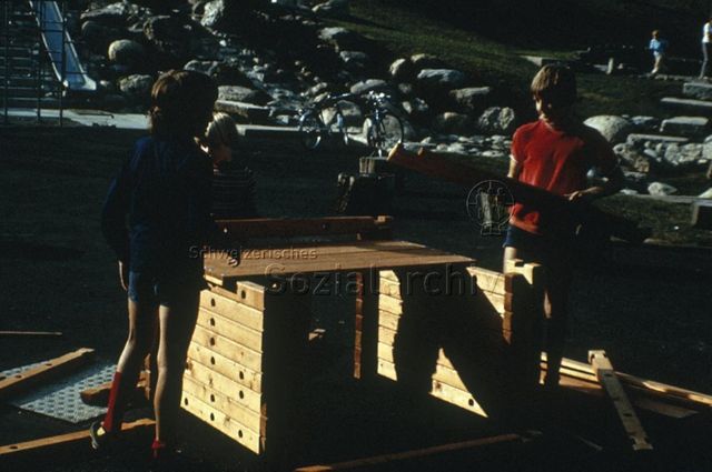 "Siedlungs- und Quartierspielplätze: Baugross Siedlungsspielplatz Adliswil ZH" - Drei Kinder am Bauen mit zusammensteckbaren Holzteilen auf dem Hartplatz; um 1970
