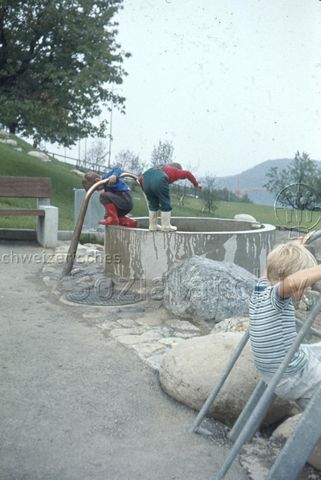 "Siedlungs- und Quartierspielplätze: Siedlungsspielplatz Adliswil ZH, 1970" - Zwei Kinder in Gummistiefeln spielen bei einem runden Brunnen, beide stehen auf dem Brunnenrand