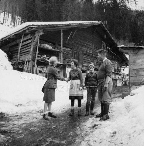 Zwei Frauen geben sich die Hand, ein Junge und ein Mann stehen daneben, hinten ein Stall/Wohnhaus; um 1965