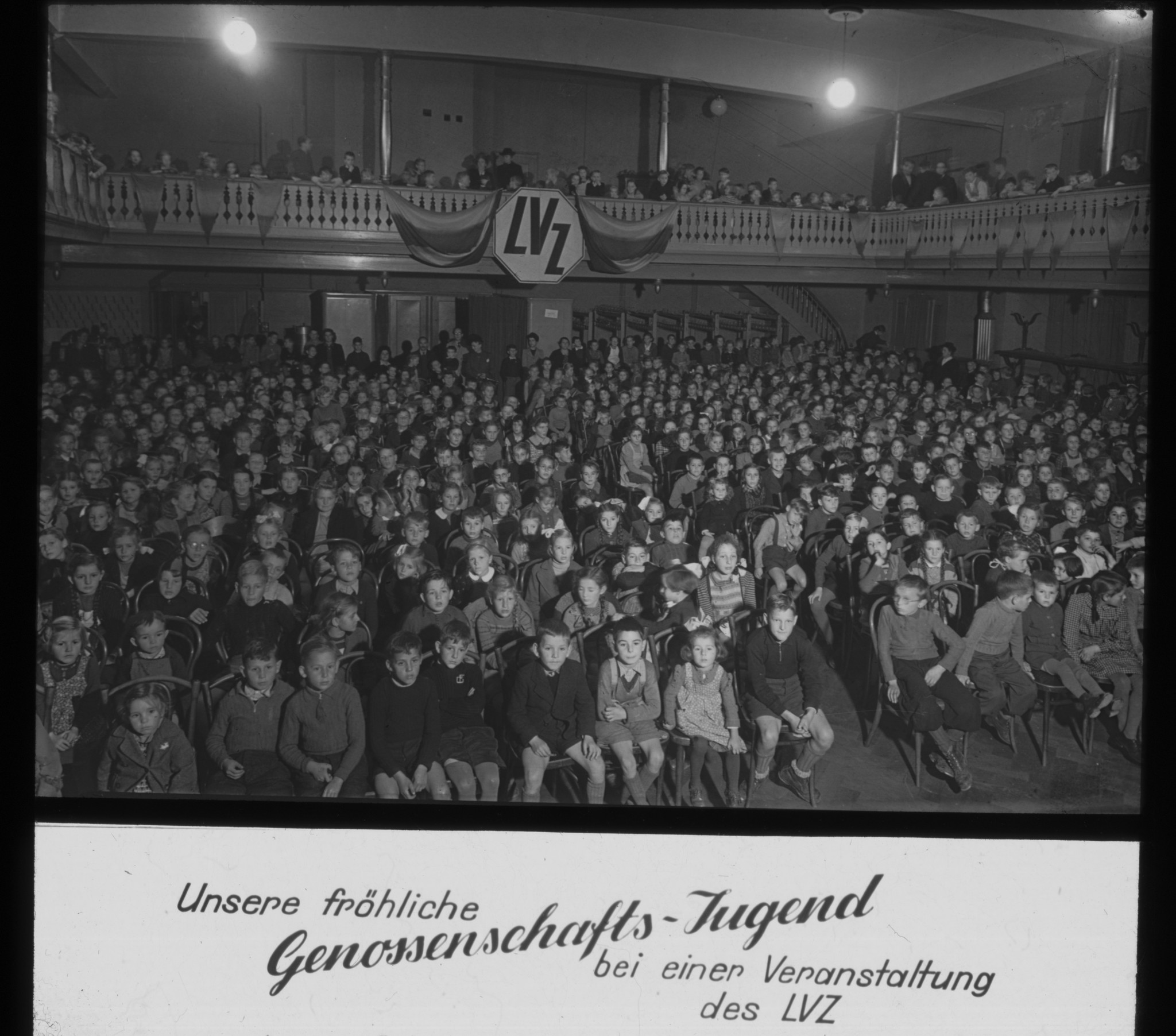 Dia: Kinder zuschauend bei einer (Heidi?)-Aufführung im Zürcher Volkshaus. Im Hintergrund aufgehängt ein LVZ-Logo, dazu Text: "Unsere fröhliche Genossenschafts-Jugend bei einer Veranstaltung des LVZ".