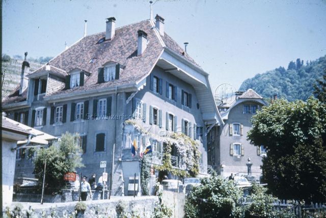 "Jugendhäuser: Gesamtansicht Maison Visinand, Montreux VD" - Aussenansicht eines grossen Hauses mit steilem Ziegeldach und einer Terrasse zu der Strasse mit Dorfbrunnen hin; um 1970