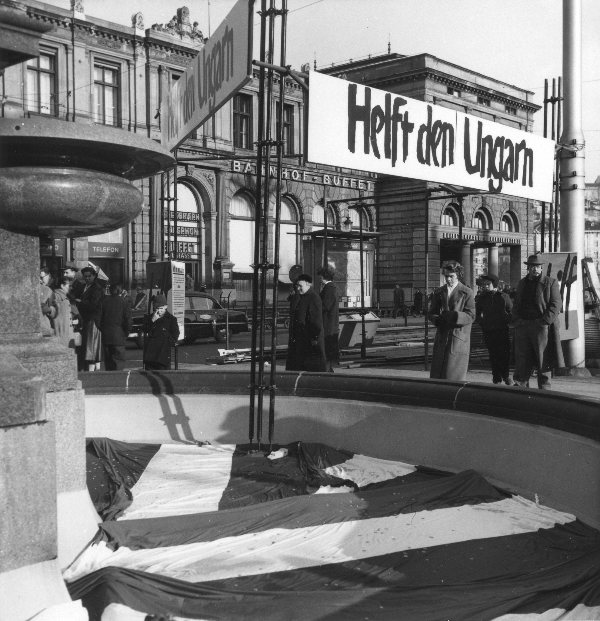 Menschen werfen Münzen in den leeren Brunnen am Bahnhofsplatz in Zürich, als Spende für Ungarn; im Hintergrund das Bahnhofsgebäude und Plakate mit der Aufschrift "Helft den Ungarn"