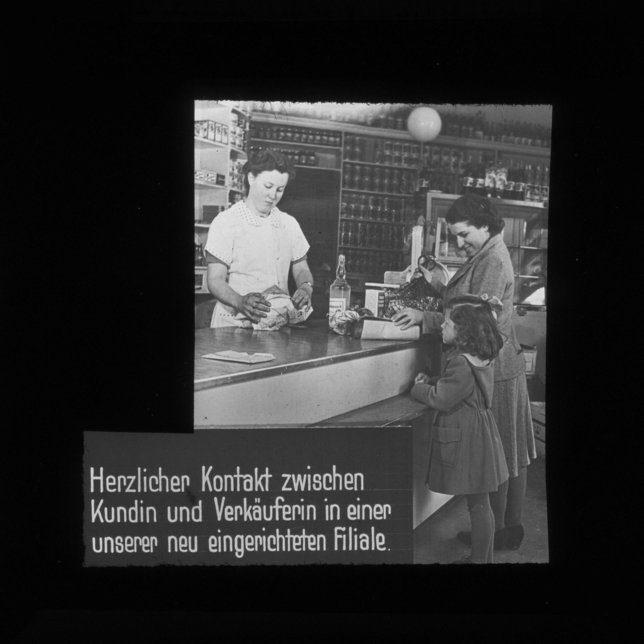Dia: Verkaufsladen, an der Theke die Verkäuferin, eine Kundin und ihre Tochter, dahinter Regale. "Herzlicher Kontakt zwischen Kundin und Verkäuferin in einer unserer neu eingerichteten Filiale."