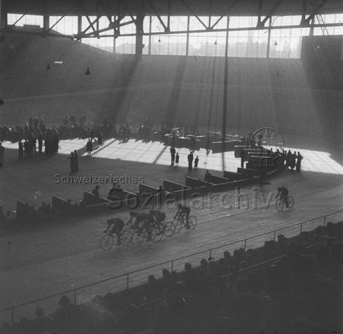 "Sport; Wettkampf: Rennbahn Oerlikon, Zürich" - Fahrradfahrer auf einer Rundbahn in einer industriell anmutenden Halle durch die Fenster strömt Sonnenlicht hinein, die Publikumsränge sind besetzt; um 1960