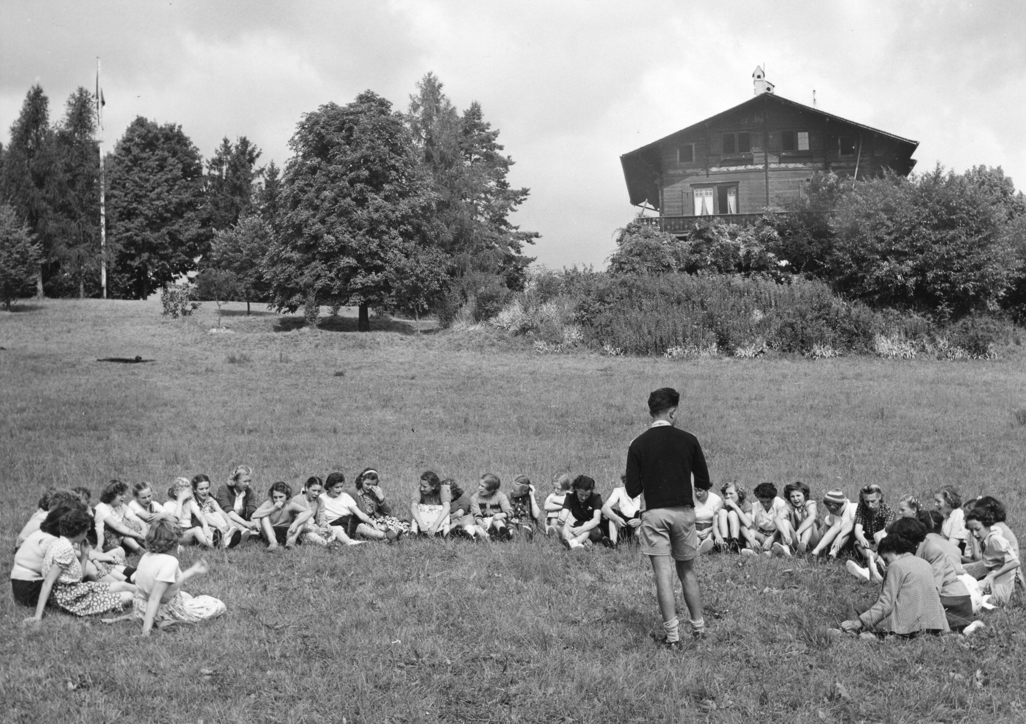 "La Fenière", Collex b. Genf, Belgierkolonie, Sommer 1950"; Kinder sitzen im Halbkreis auf einer Wiese und hören einem Betreuer zu