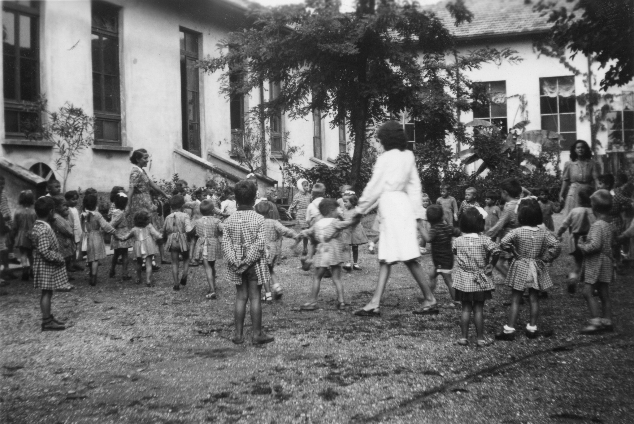 "Vormittags zwischen 11 und 12 Uhr im Garten. (Lehrerinnen: v.l.n.r.: Adeline Crameri, Amelia Cagna, Flora Franzoso)"; Kinder tanzen im Kreis