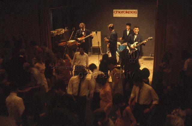 "Diskothek: Disco im FZA" - Vier junge, in Anzüge gekleidete Musiker mit Schild "cherokees" auf der Bühne, die Leute tanzen; um 1965