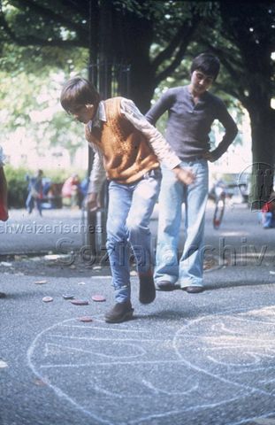 "Hüpfspiele" - Zwei Jungen beim Spielen auf einem aus Kreide aufgemalten Feld; um 1975