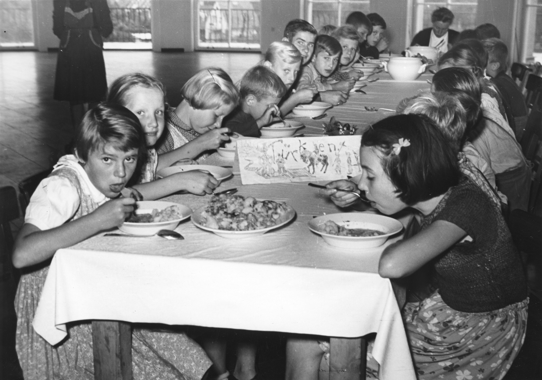 "Sommer 1953 - Hörnwald: Ferien für Flüchtlingskinder mit Hilfe des Schweizerischen Arbeiterhilfswerkes und der Schweizer Europahilfe. Volkshilfe Wien". Kinder sitzen essend an einem Tisch