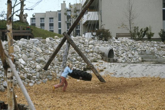 "Siedlungs- und Quartierspielplätze: Siedlungsspielplatz Widen/ Mutschellen AG" - Mädchen spielt mit einem an Ketten befestigten Gummireifen auf einem Spielplatz im Wohnquartier; um 1970