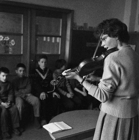Lehrerin spielt Geige, die Schüler sitzen in einer Reihe; um 1970