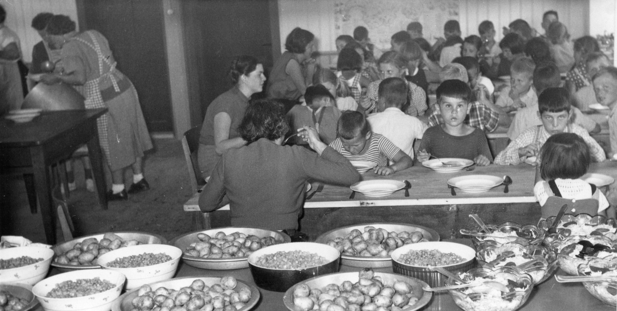 Kinderhilfe Schweiz: ein Saal mit Kindern und Heimleiterinnen essend, im Vordergrund das Buffet mit Kartoffeln, Erbsen und Salat
