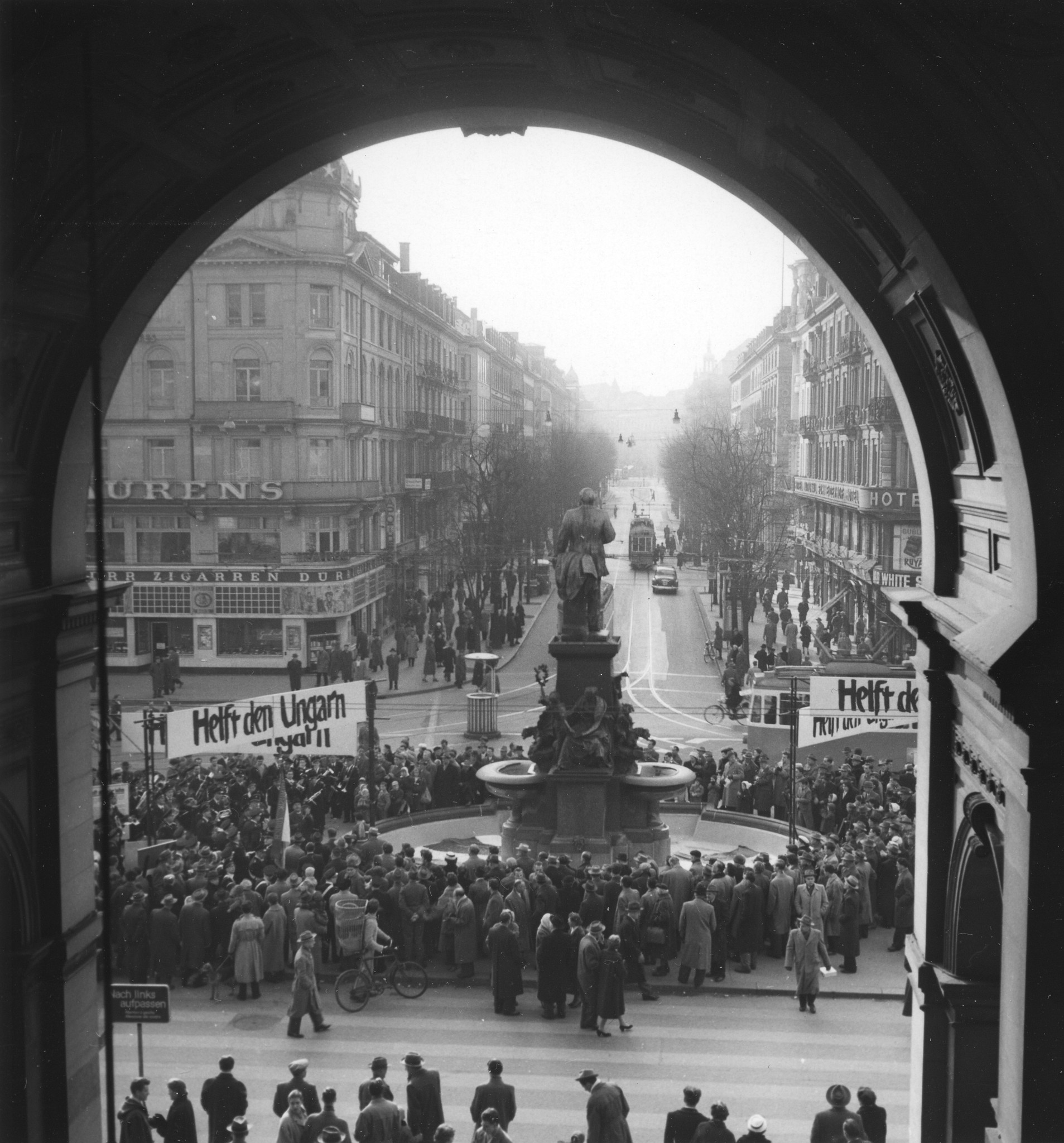 Blick durch den Torbogen auf den Bahnhofsplatz und die Bahnhofsstrasse in Zürich; Menschen mit den Schildern "Helft den Ungarn" stehen auf dem Bahnhofplatz und demonstrieren