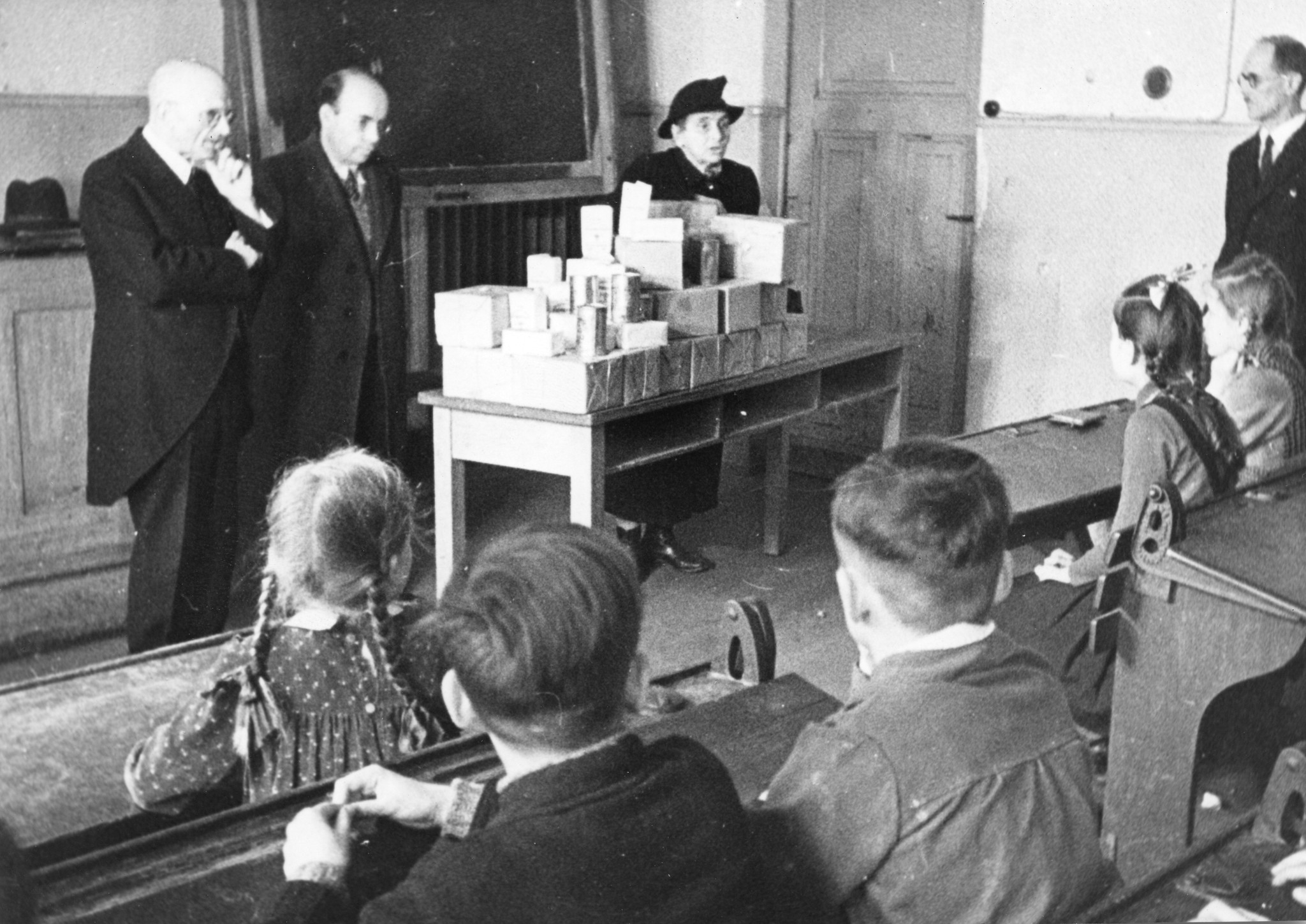 "März 1948: Verteilung von Lebensmittelpaketen am die Schüler der Gemeinschaftsschule Düsseldorf"; eine Frau hinter einem Tisch mit Lebensmitteln stehend und mit mit den Kindern sprechen, drei weitere Männer hören zu