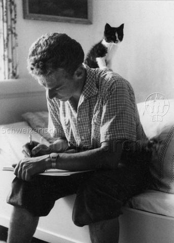 PraktikantInnenhilfe - Praktikant sitzt auf dem Bett und schreibt in ein Buch, auf seinem Rücken sitzt eine Katze; um 1965