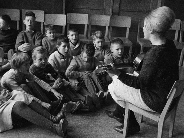 Schule - Kinder beim Musikunterricht, Lehrerin begleitet die singenden SchülerInnen auf der Gitarre; um 1965