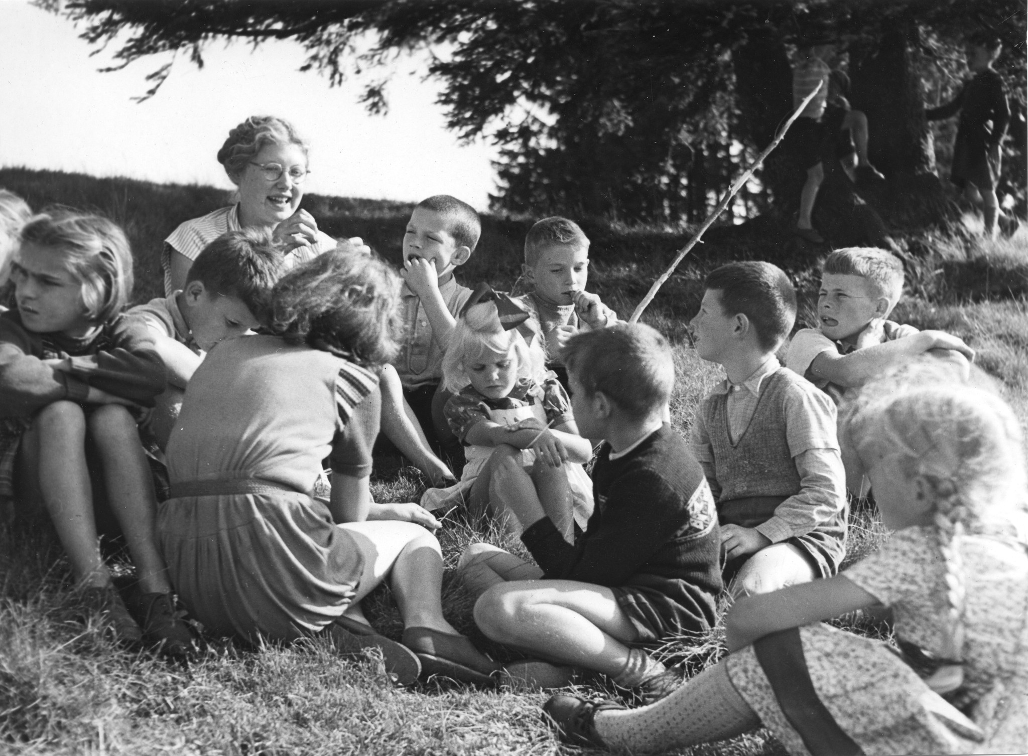 Ferienkolonie Landmark: Kinder sitzend mit einer Betreuerin auf einer Wiese und hören ihr zu