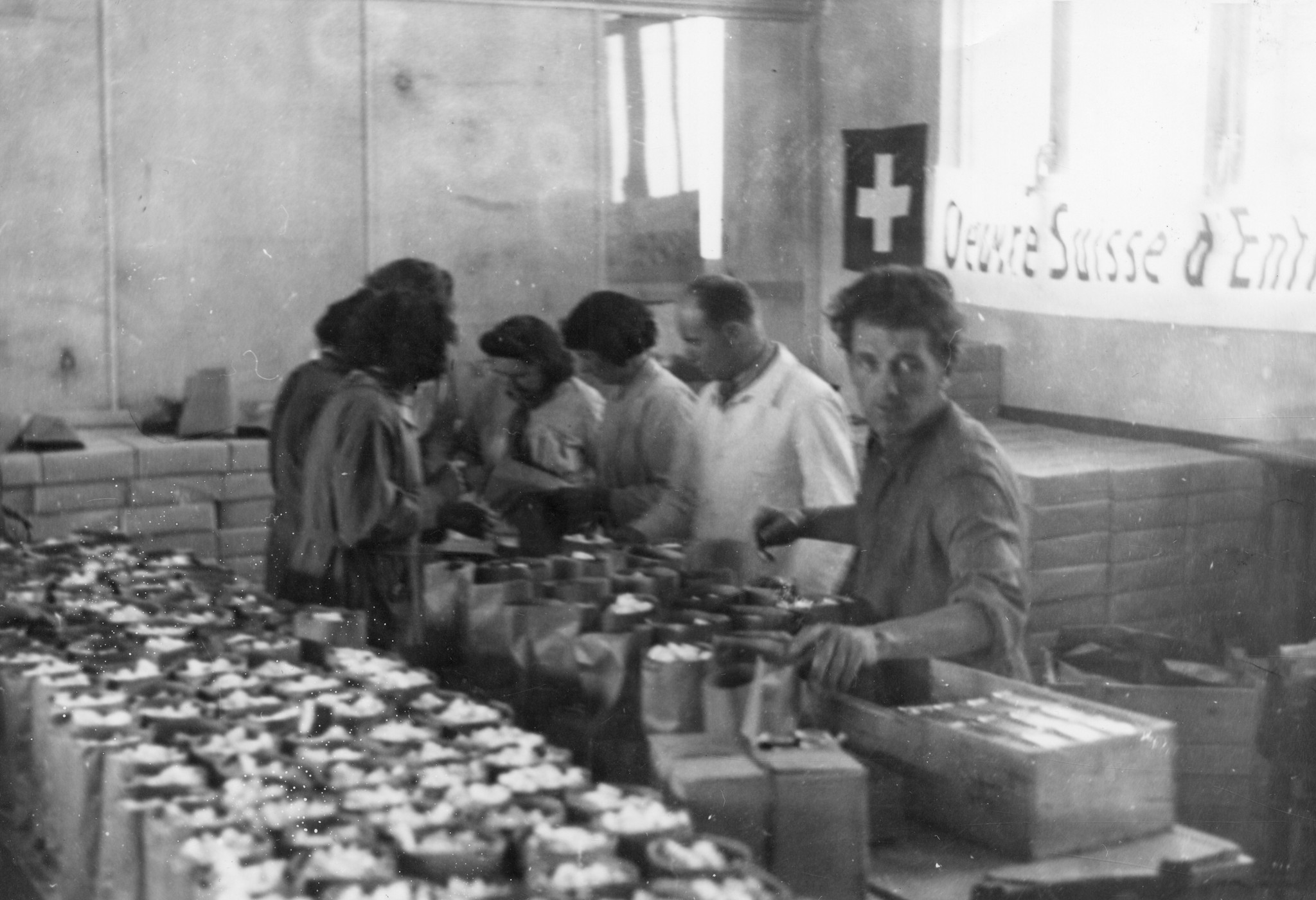 "Lyon, Oktober 1944. Seit Montag morgen werden Tüten gemacht, und die Füllung der Tüten wird vorbereitet." Helfer beim Vorbereiten von Rationen mit Zückerwürfeln, Transparent: "Oeuvre Suisse d'Entraide Ouvrière"