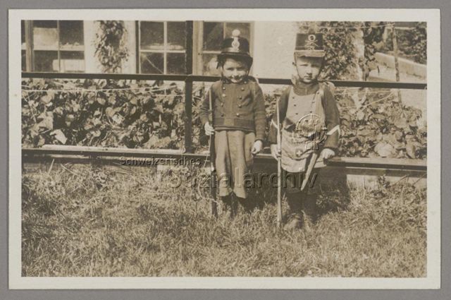 Zwei Knaben im Garten in Militärverkleidung mit Infanterieuniform, Holzgewehr und (Füsilier-)Tschako, um 1910