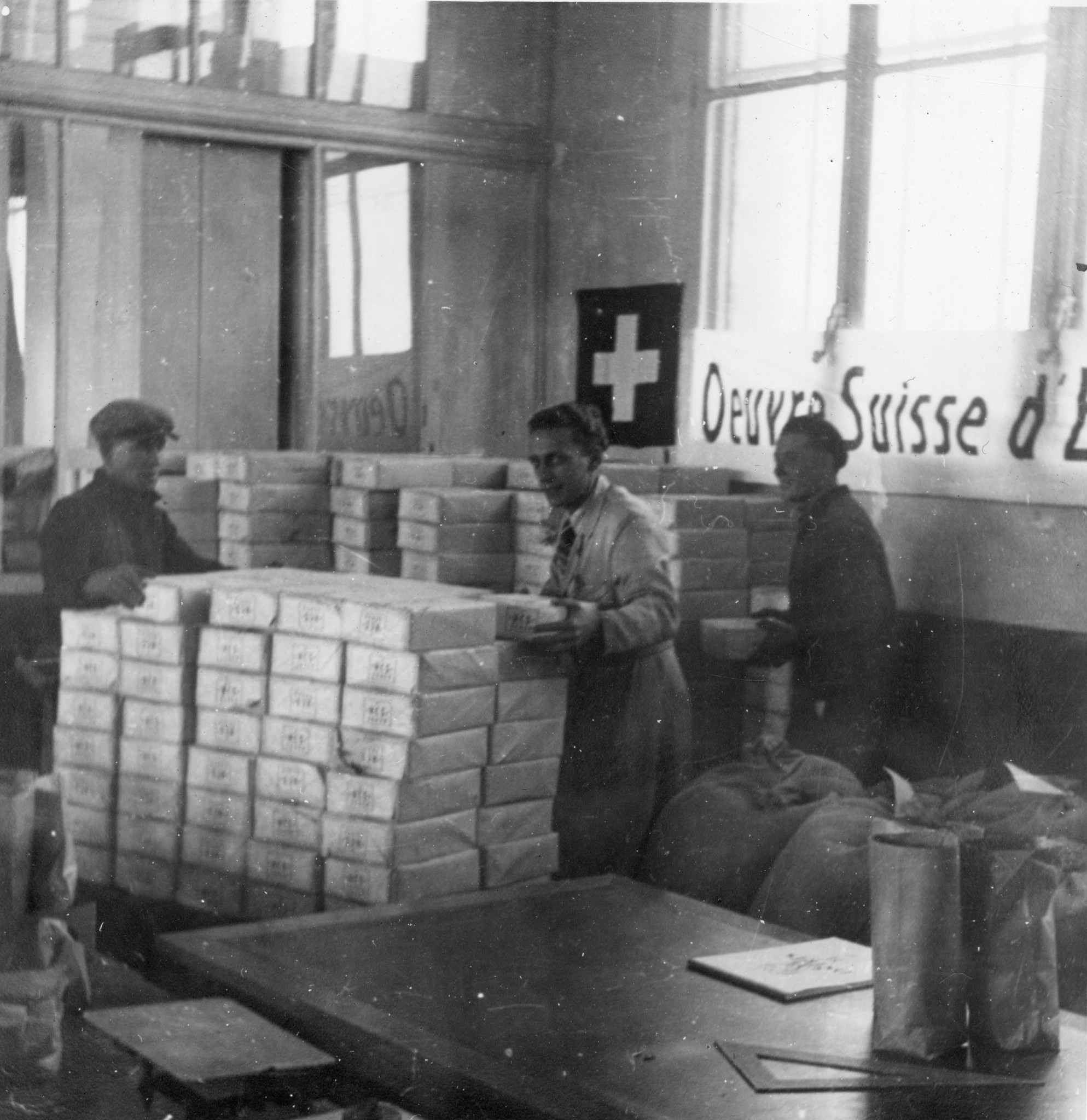 "Lyon, Oktober 1944. Das Lebensmittelmagazin wird sofort eingerichtet am Tage der Ankunft. Es ist Freitag nachmitag. Am Samstag nachmittag ist die Fabrik geschlossen." Transparent des SAH im Hintergrund: "Oeuvre suisse d'entraide ouvrière"
