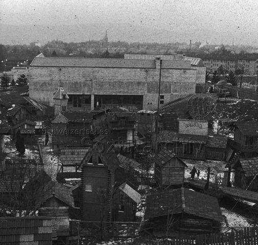 "Freizeitanlagen, Gemeinschaftszentren: Robinsonspielplatz Heuried, Zürich" - Blick auf die Anlage mit zahlreichen Spielhäusern, eines mit der Aufschrift "Saloon" vor einem Betongebäude; um 1960