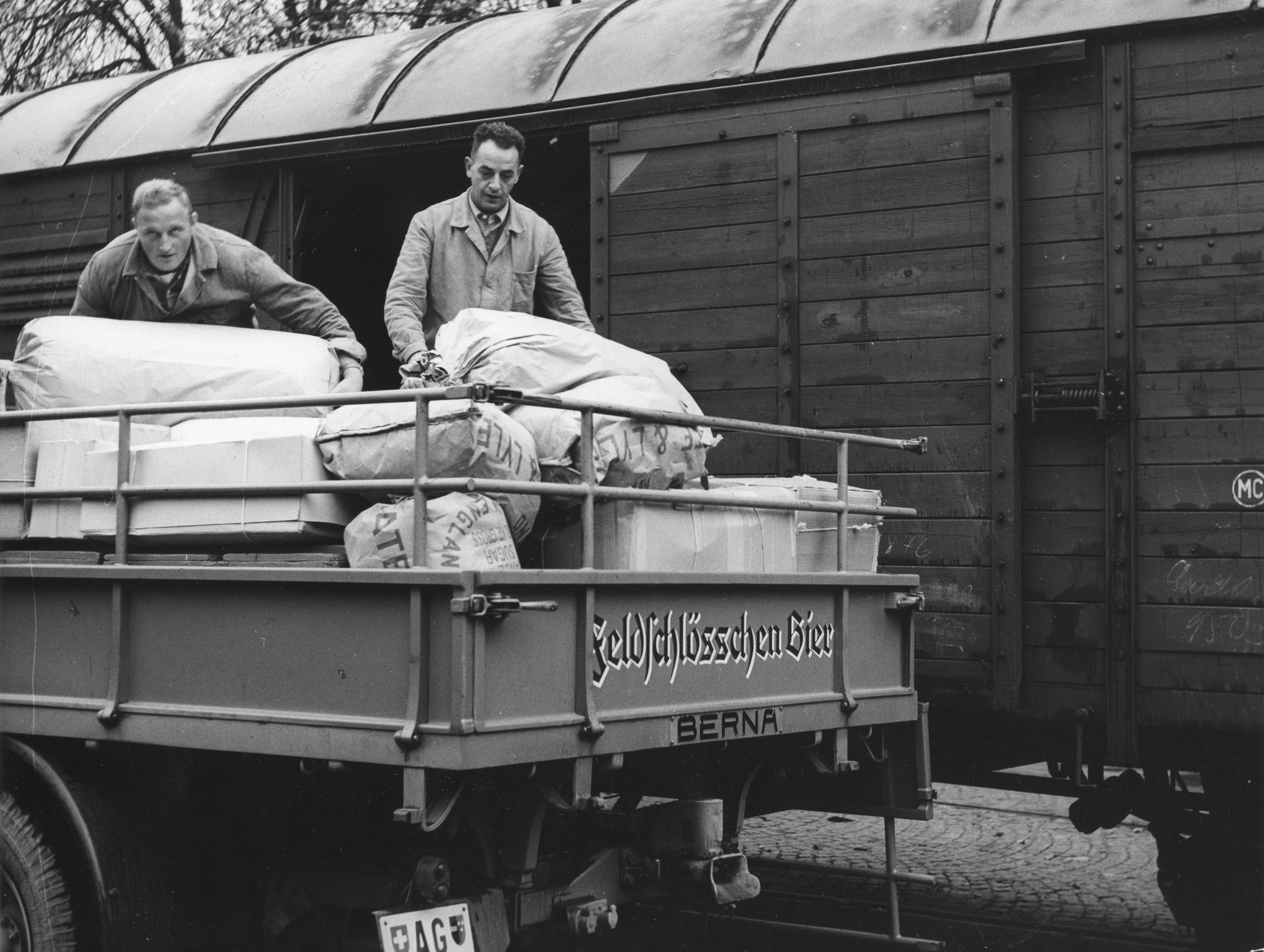Zwei Männer heben Säcke von einem Lastwagen mit der Aufschrift "Feldschlösschen Bier"
