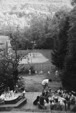 Bosco della Bella, Feriendorf für kinderreiche Familien - Kinder und Erwachsene sitzen um zwei Tische, im Hintergrund wird Fussball und Federball gespielt; um 1975