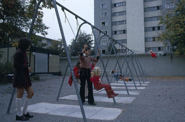 "Siedlungs- und Quartierspielplätze: Siedlungsspielplatz Hirzenbach Schwamendingen, Zürich" - Kinder schaukeln auf einem, von einer Mauer umgebenen, Platz mit Schottersteinen, im Hintergrund sieht man einen Wohnblock; um 1970