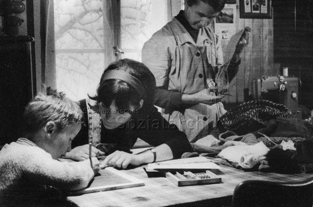 "Praktikantinnenhilfe" - Praktikantin hilft Jungen beim Schreiben auf einer Tafel; um 1965
