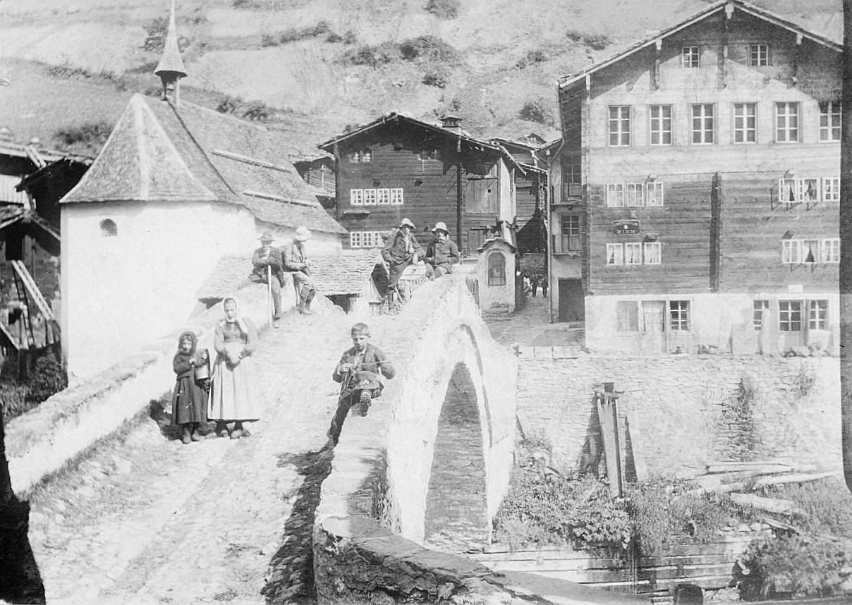 Am Sonntagmorgen in Binn (Binntal, Wallis): Dorfbewohner (vermutlich) bei der Brücke, im Hintergrund ist die Dorfkirche sichtbar - G