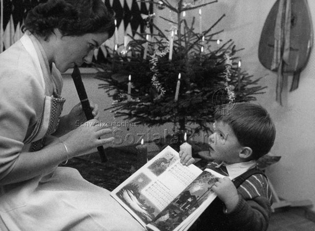 Weihnachtsfest - Junge Frau spielt ein Lied auf der Flöte, ein kleiner Junge hält ihr das Notenbuch, im Hintergrund brennen Kerzen an einem Christbaum; um 1965
