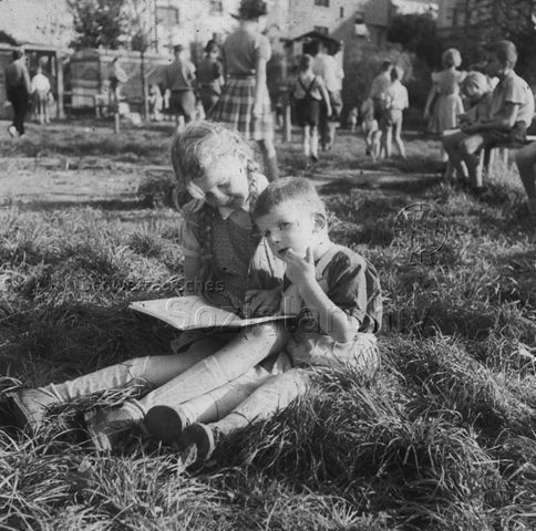 "Freizeitanlagen, Gemeinschaftszentren: FZA Wipkingen, Zürich" - Kinder auf einer Spielwiese, ein Mädchen liest einem kleineren Jungen aus einem Buch vor; um 1960