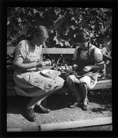 Diaserie zum Thema Mädchen; zwei Mädchen sitzen auf einer Holzbank im Freien und rüsten Äpfel und Karotten; um 1940