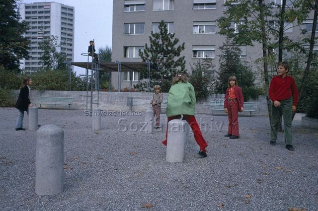 "Siedlungs- und Quartierspielplätze: Siedlungsspielplatz Hirzenbach Schwamendingen, Zürich" - Kinder spielen auf einem Platz mit Schottersteinen, ein Mädchen hüpft über einen steinerenes Böckli; um 1970