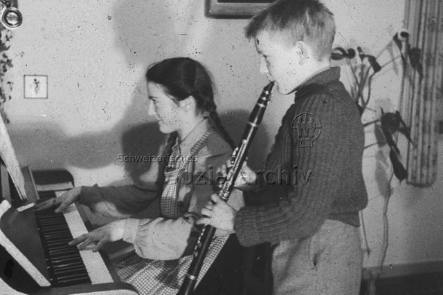 "Musik: Hausmusik" - Ein Junge mit einer Klarinette und ein Mädchen am Klavier, das Mädchen hat lange Zöpfe und trägt eine Schürze, um 1950
