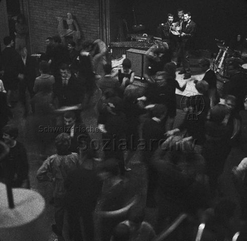 "Freizeitanlagen, Gemeinschaftszentren: Tanzabend in der FZA Bachwiesen, Zürich" - Drei junge Musiker in Anzügen auf der Bühne, Paare am Tanzen; um 1965