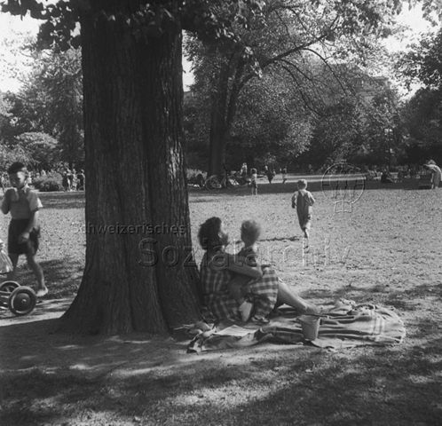 "Quartierspielwiese: Die Bäckeranlage, Zürich" - Parkanlage mit grossen Bäumen, eine Mutter sitzt mit ihrem Kind auf einer Picknickdecke im Schatten, ein Kind rennt über die Wiese; um 1960