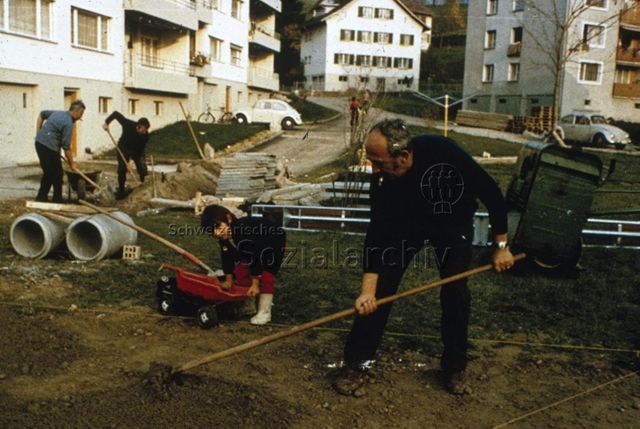 "Siedlungs- und Quartierspielplätze: Fronarbeit Siedlungsspielplatz, Ebnat-Kappel SG" - Baustelle des Kinderspielplatzes auf einer grossen Wiese, ein Mann ebnet den Boden, ein Kind mit seinem Lastwagen; um 1970