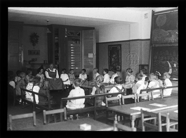 Diaserie zum Thema Kindergarten; Kinder und eine Frau sitzen in einem Schulzimmer auf Holzbänken im Kreis; um 1930