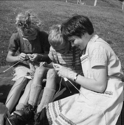Praktikantinnenhilfe - Zwei Kinder und Praktikantin sitzen auf einer Wiese und stricken; um 1965