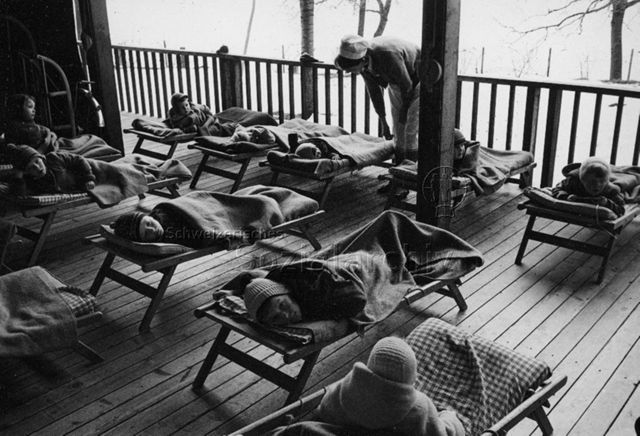 "Kinderheilstätte [Davos], Liegekur" - Kinder liegen, gut zugedeckt und eingekleidet, draussen auf Feldbetten; um 1970