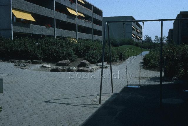 "Siedlungs- und Quartierspielplätze: Augarten Rheinfelden AG" - Kinderschaukel an einem Gehweg zwischen Büschen und Wiesen in einem Quartier mit Wohnblöcken; um 1970