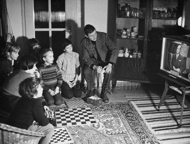 "Schachsendereihe - (...) 'Im Banne der 64 Felder' ist ein Einführungskurs in das Schachspiel. (...) Die Zuschauer haben die Möglichkeit, zu Hause vor dem Bildschirm die Spielvorgänge auf ihrem eigenen Schachbrett mitzuspielen (...)" - Eine Familie schaut gebannt in den Fernseher; 1962