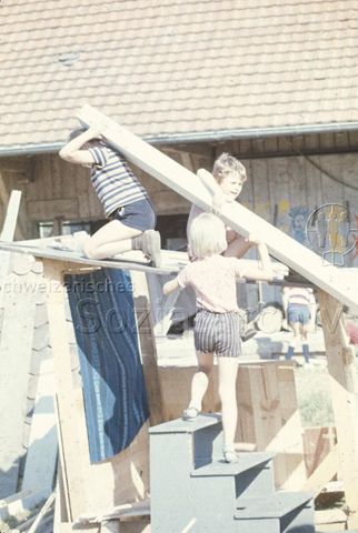 "Bauspielplatz Binningen" - Drei Kinder heben einen Holzbalken auf ihre Hütte; um 1975