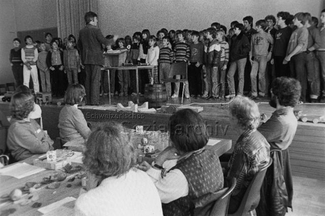 Kinder beim Singen an einer schulischen Veranstaltung, die Eltern sitzen im Publikum; um 1980