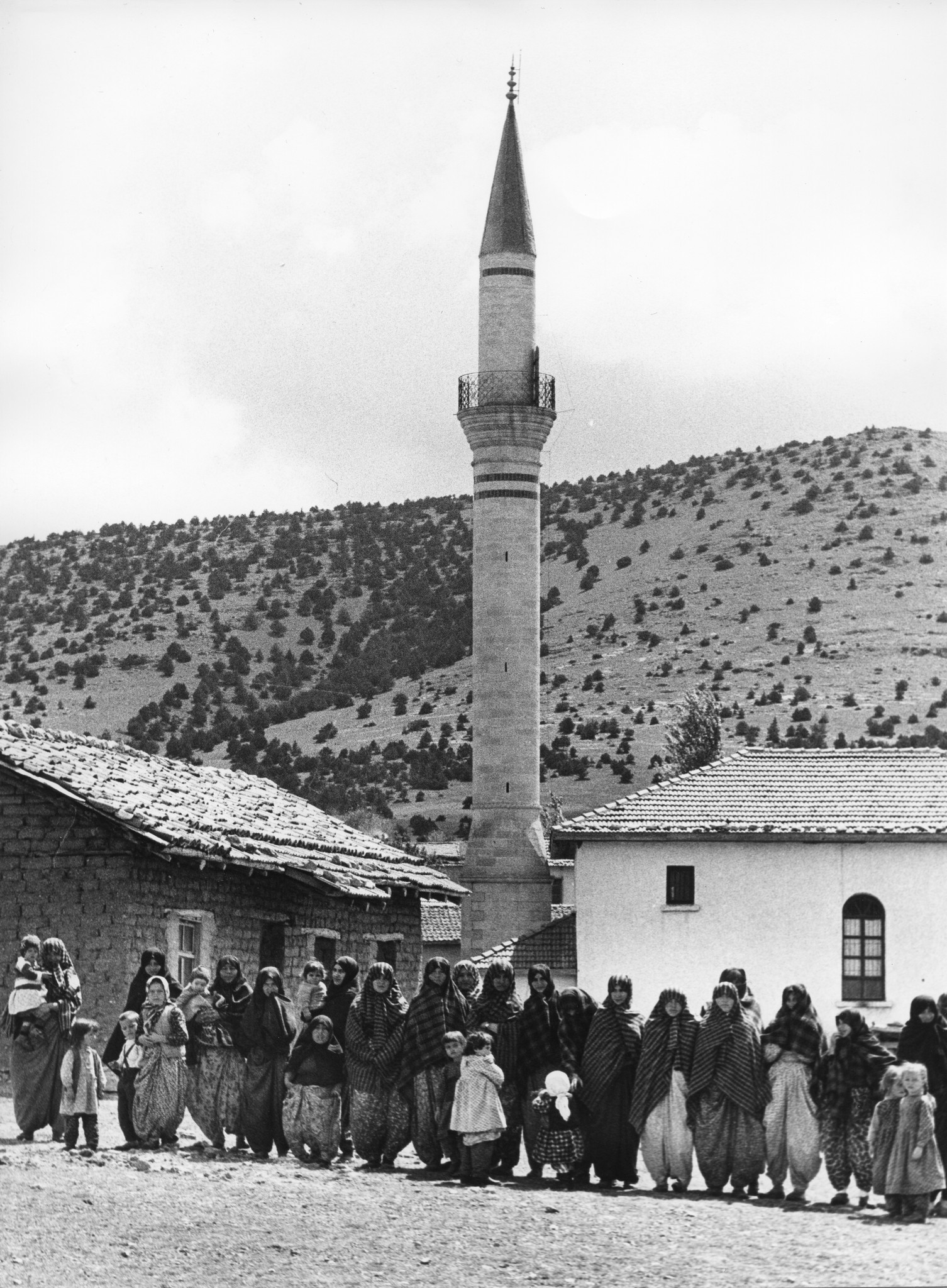 In einem türkischen Dorf stehen Frauen und Kinder in einer Reihe, im Hintergrund eine Moschee mit Minarett
