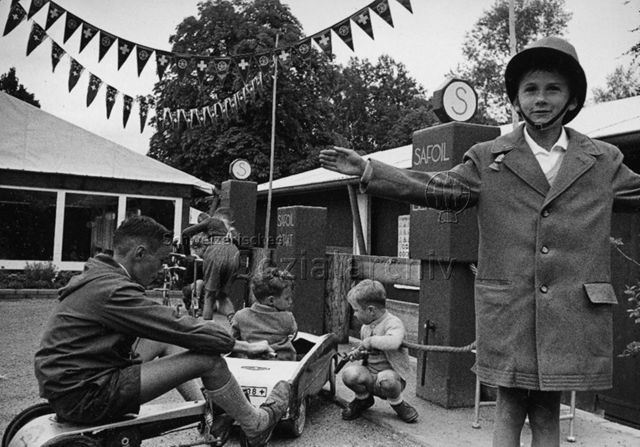 Jungen beim Spielen mit Tretautos an Spiel-Tankstelle, ein Junge im Vordergrund regelt den Verkehr; um 1965