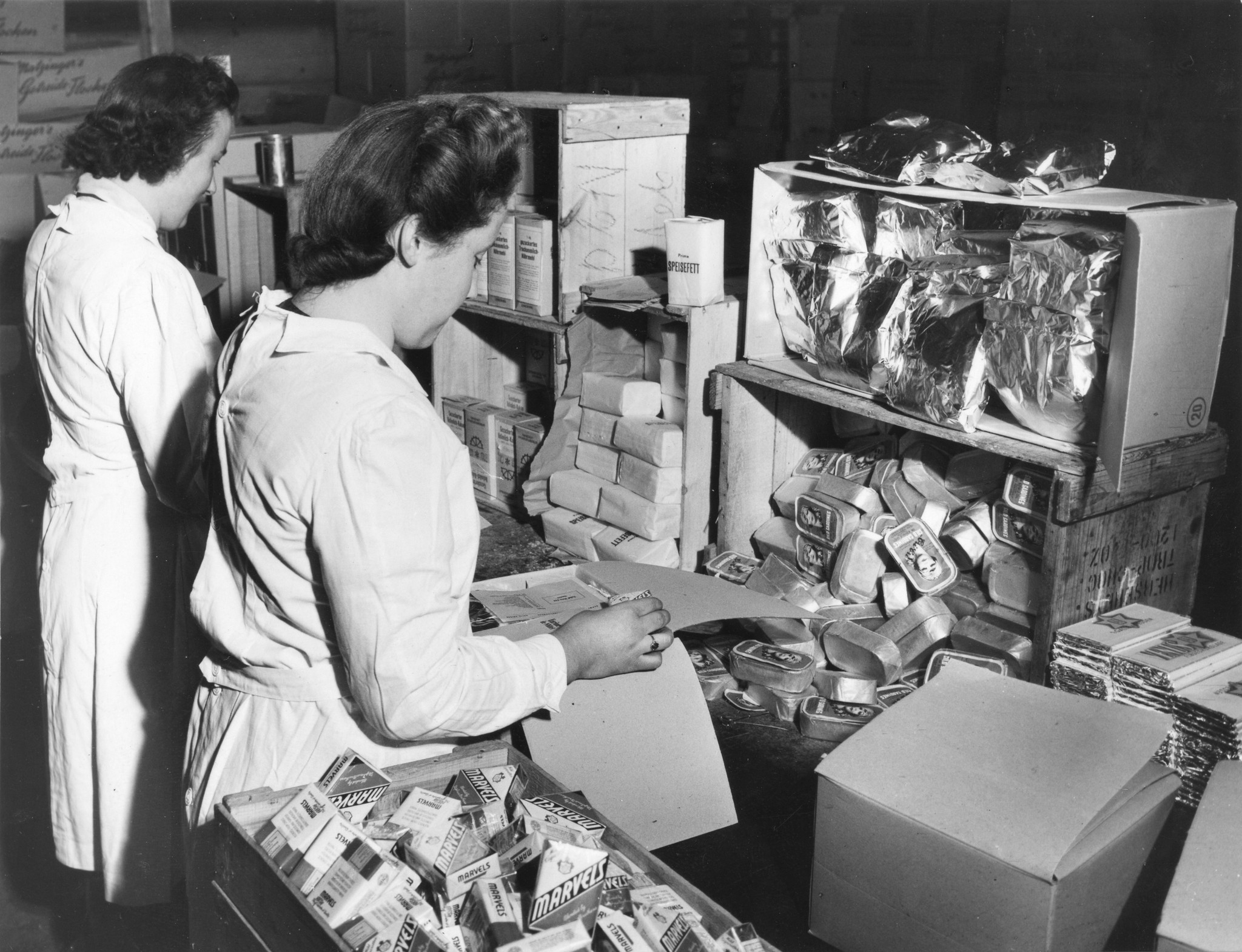 "Colis werden zusammengestellt", zwei Frauen bei der Vorbereitung von Lebensmittelpaketen (Sardinenbüchsen, "Marvels", Schokolade, Speisefett, Trockenmilch...), um 1950 (Nachkriegszeit)