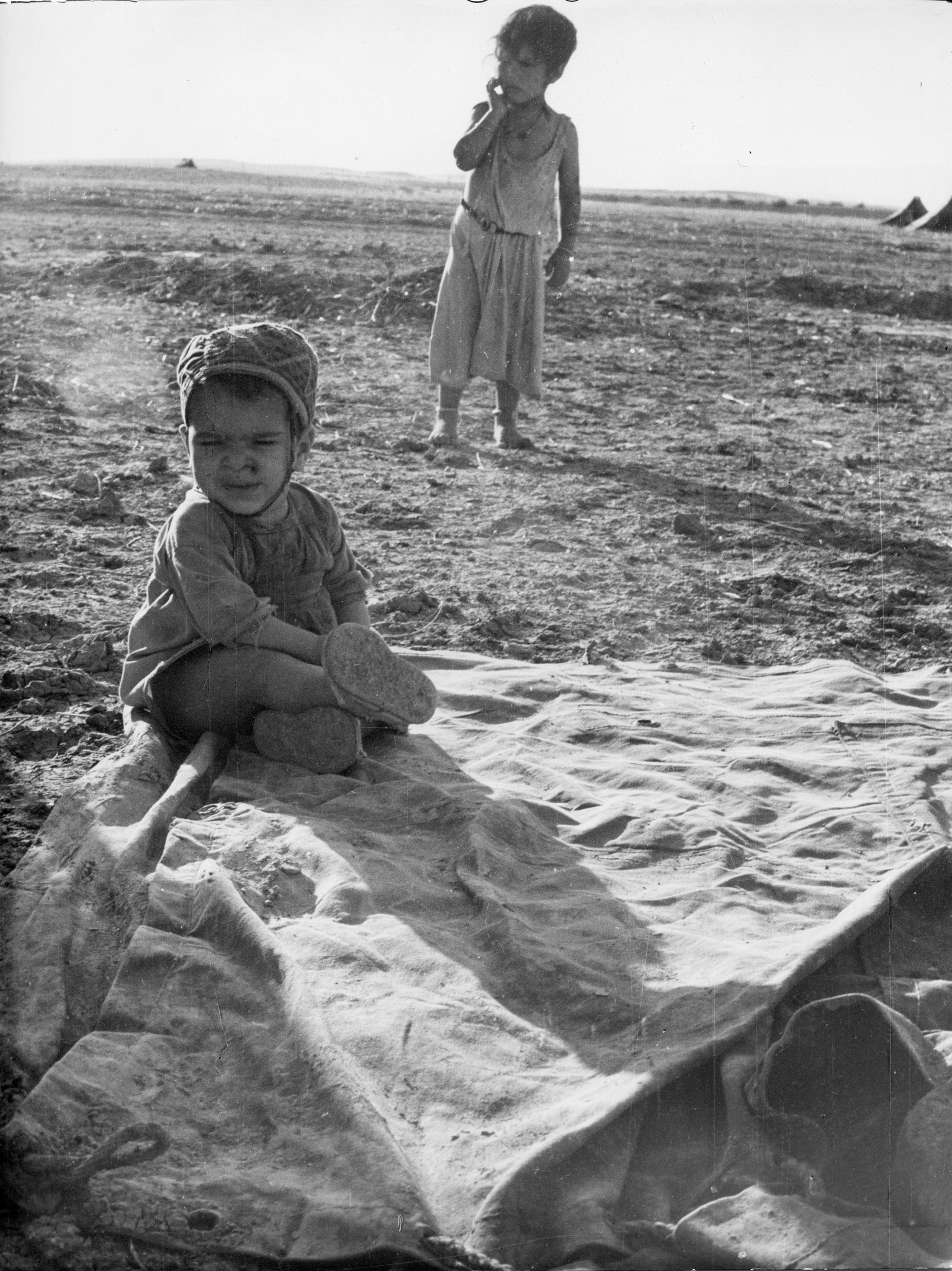 Ein Kleinkind sitzt im Sand, ein älteres Kind steht weiter hinten